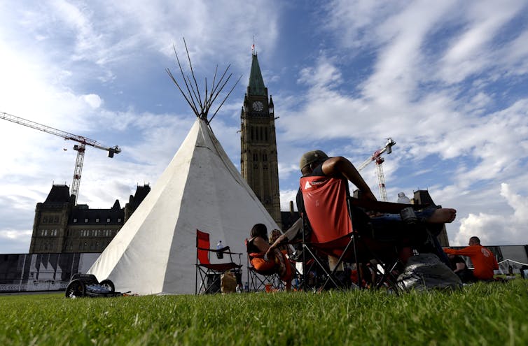 People seen in chairs in front of a teepee and Parliament Hill.