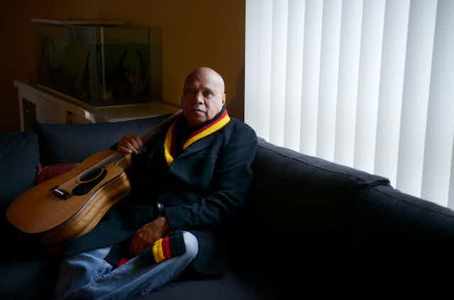 Uncle Archie roach sits on a coach with his guitar and a scarf adorned with colours of the Aboriginal flag; black red and yellow.