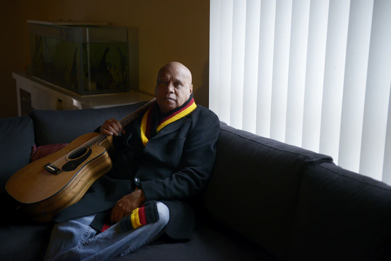 Uncle Archie roach sits on a coach with his guitar and a scarf adorned with colours of the Aboriginal flag; black red and yellow.