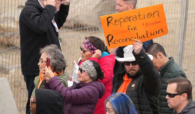 A man in sunglasses is seen with an orange sign that says 'Reparation for Reconciliation, $30 milliion.'  