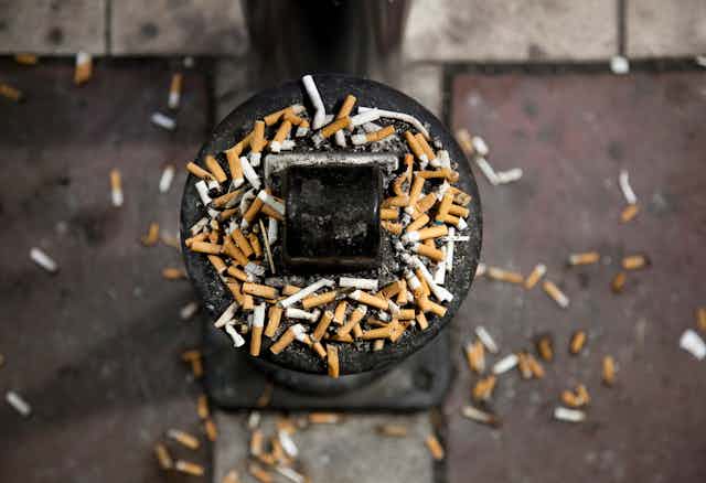 Cigarette butts spilling out of an outdoor ashtray and onto the ground around it.