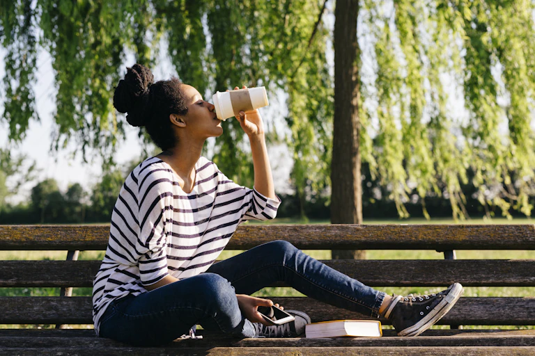 A young woman wearing casual apparel sitting on a park bench drinking coffee from a to-go cup.