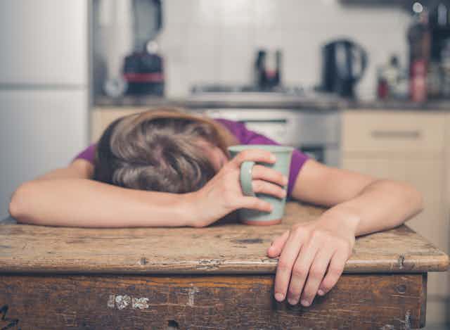 Person slumped over table asleep, clutching coffee mug