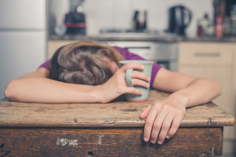 Person slumped over table asleep, clutching coffee mug