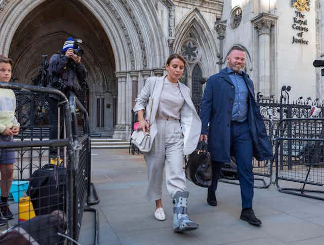 Coleen Rooney, in light grey trousers, top and jacket, wearing a medical boot on her left foot, walks out of the high court next to her husband Wayne Rooney