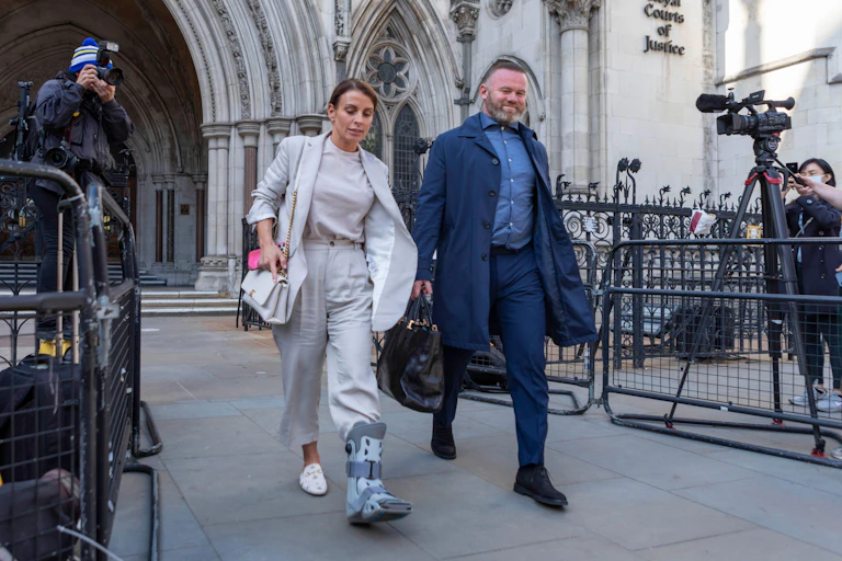 Coleen Rooney, in light grey trousers, top and jacket, wearing a medical boot on her left foot, walks out of the high court next to her husband Wayne Rooney
