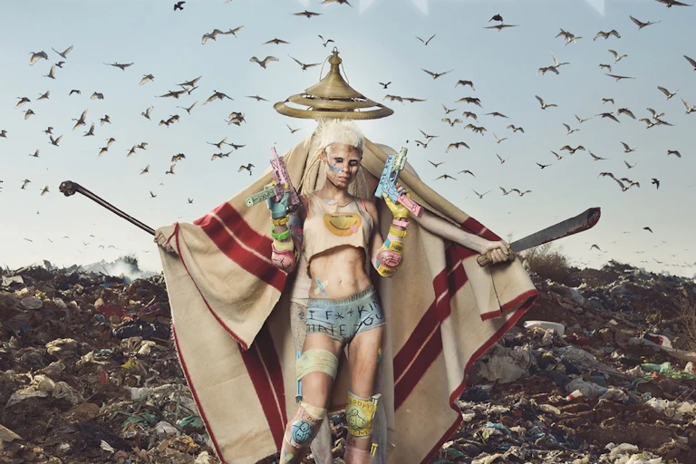 An image of a woman with short blonde hair holding guns in either hand in front of a figure in a Basotho hat and Lesotho blanket, a panga and a club in his outstretched hands.
