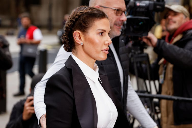 Side view of Rebekah Vardy in a white shirt and black suit jacket, with her hair neatly braided into a bun, arriving at court