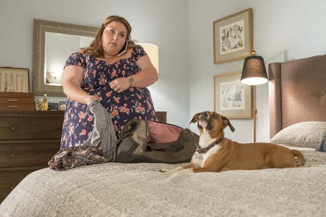 Una mujer con sobrepeso habla por teléfono móvil mientras prepara una maleta en la habitación con un perro sobre la cama.