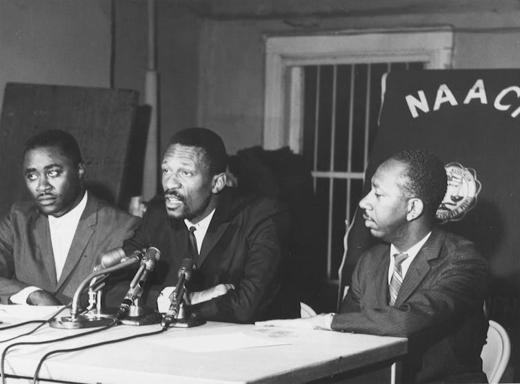 Con una pancarta de NAACP detrás de ellos, dos hombres negros se sientan en una mesa mientras el tercero habla por un micrófono.