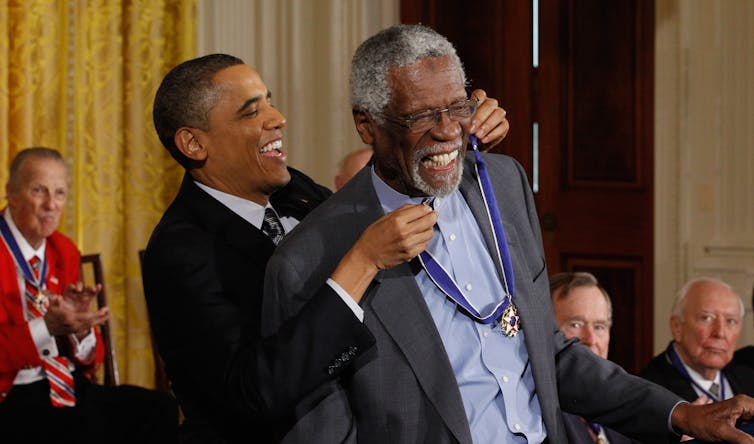 A black man wearing a dark blue suit is tying a ribbon around the neck of another black man.