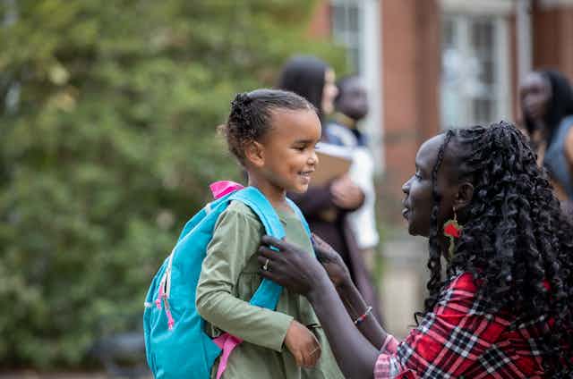 A young girl with an aqua and pink backpack is smiling as she faces her mother, who is eye level and adjusting her backpack.