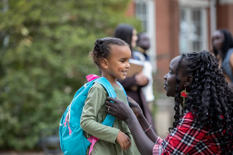 A young girl with an aqua and pink backpack is smiling as she faces her mother, who is eye level and adjusting her backpack.