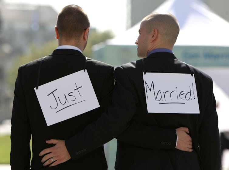 Two men wearing suits stand with their backs to the camera and signs that say Just Married on their backs.