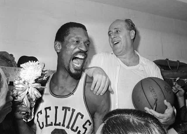 Two men, one in a basketball jersy and the other in a white shirt and undershirt, holding a basketball, laugh and celebrate.