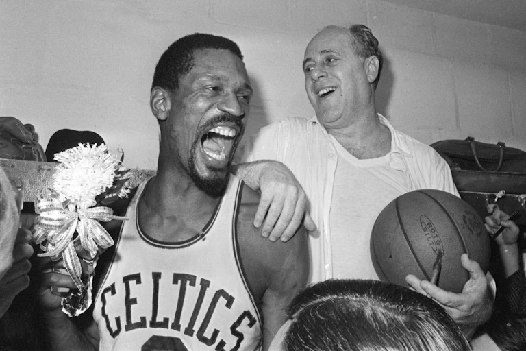 Two men, one in a basketball jersy and the other in a white shirt and undershirt, holding a basketball, laugh and celebrate.