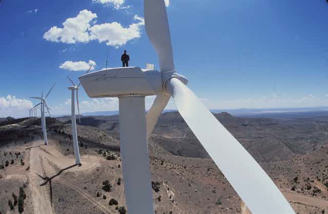 A man stands a top a wind turbine with a row of turbines behind him on a ridge in dry scrub landscape
