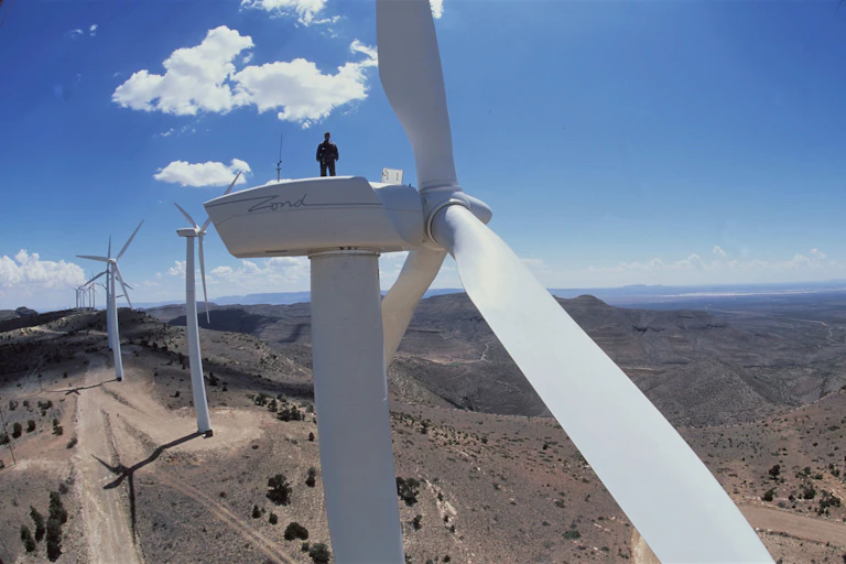 A man stands a top a wind turbine with a row of turbines behind him on a ridge in dry scrub landscape