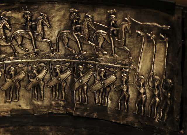 mounted cavalry and soldiers holding shields face three figures playing boar-headed carnyxes