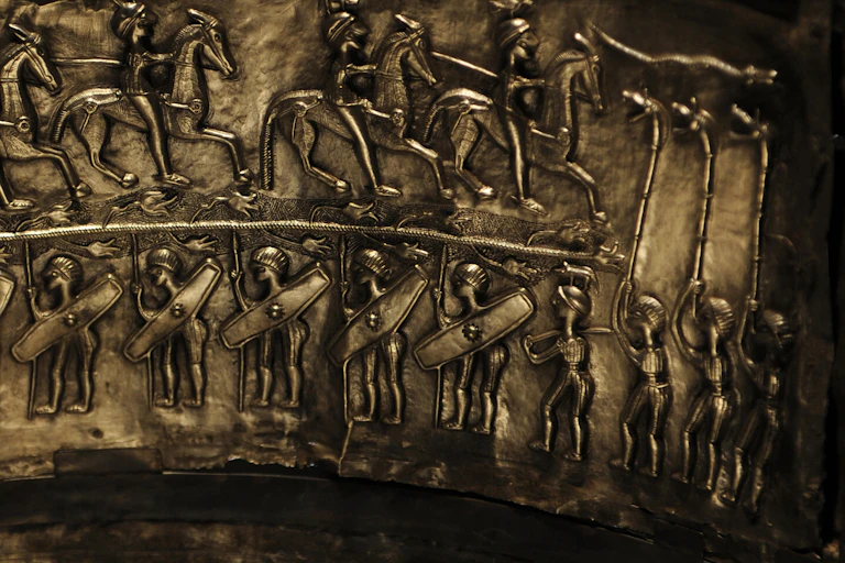 mounted cavalry and soldiers holding shields face three figures playing boar-headed carnyxes