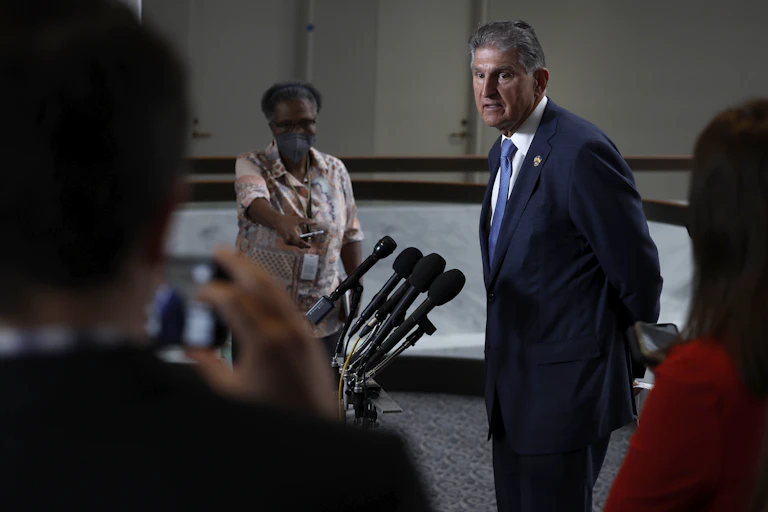 A man in a blue suit, white shirt and blue tie speaks to reporters in front of a bank of microphones.