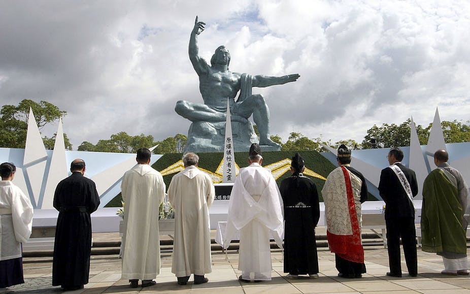 Nine men, many in robes, stand with their backs to the camera in front of a statue of a muscular man pointing to the sky.