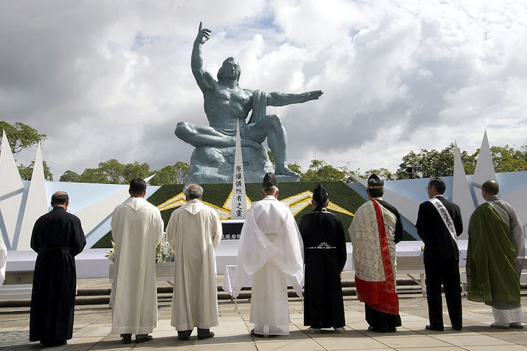 Nine men, many in robes, stand with their backs to the camera in front of a statue of a muscular man pointing to the sky.