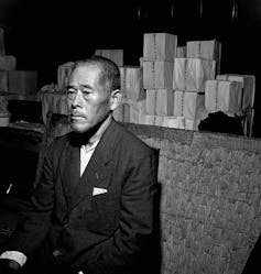 A black and white photo shows a man looking sad sitting in front of small boxes with Japanese script.