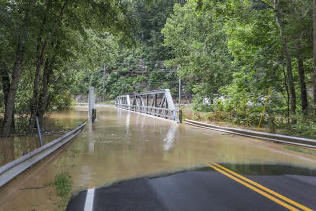 A road leads into brown waters overtopping a bridge, surrounded by trees.