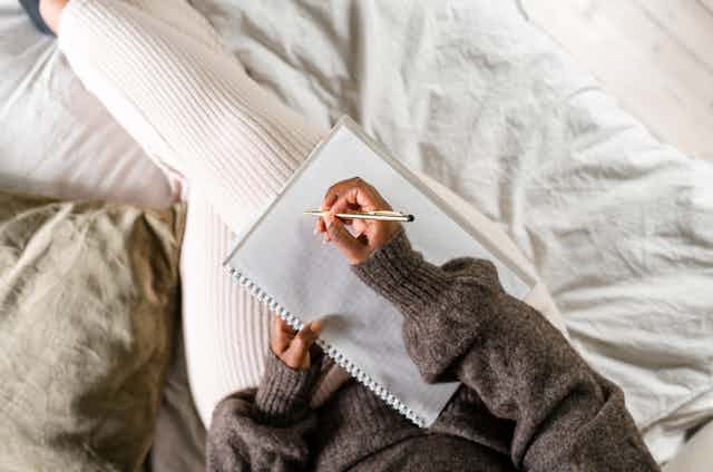 A woman sits on her bed and uses a notepad to write.