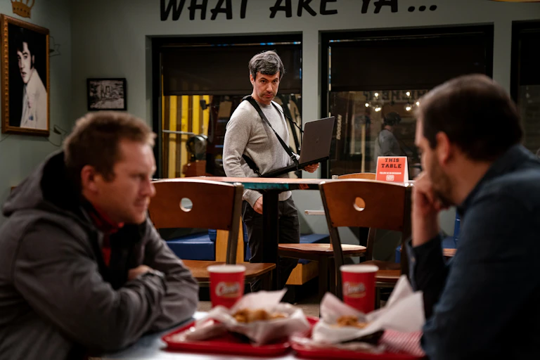 A man has a laptop hanging from his neck, as he watches a conversation.