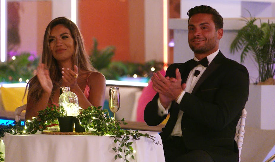 Ekin-Su and Davide in formal clothes, sitting at a table and applauding