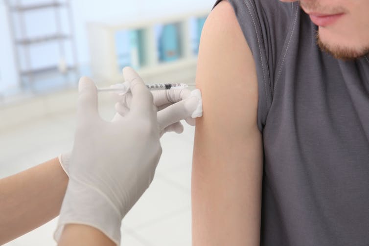 A young man receives a vaccine from a healthcare worker. The worker is wearing white surgical gloves to administer the needle.