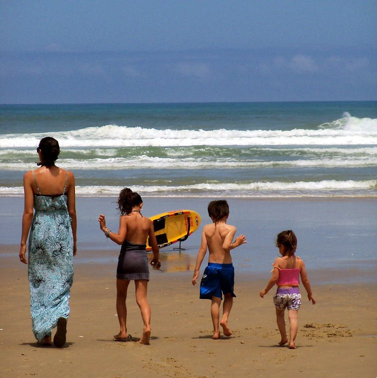 Una mujer y tres niños en bañador caminan hacia la orilla en una playa.