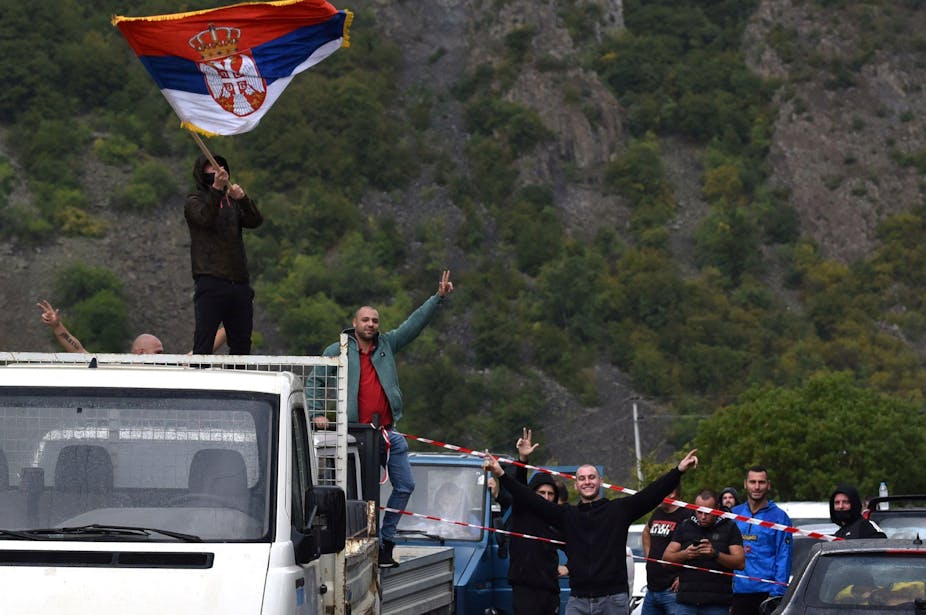 A man standing on a truck holding up a Serbian flag.