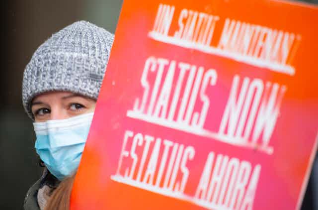A woman in a wool hat wearing a mask carries a sign that reads Status Now.