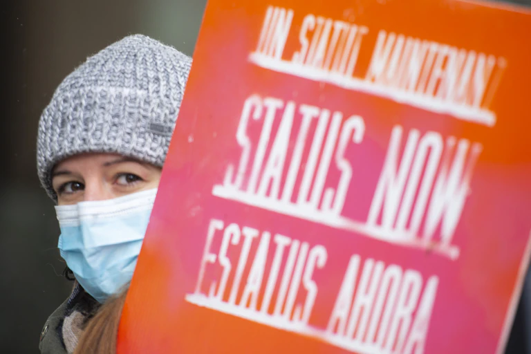 A woman in a wool hat wearing a mask carries a sign that reads Status Now.