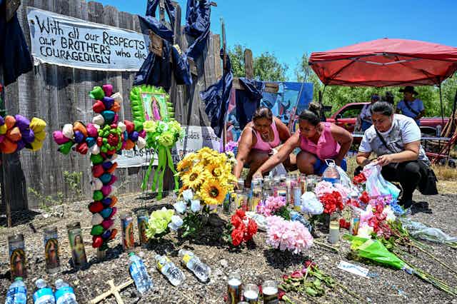 People place flowers and candles near a fence, alongside colorful homemade crosses