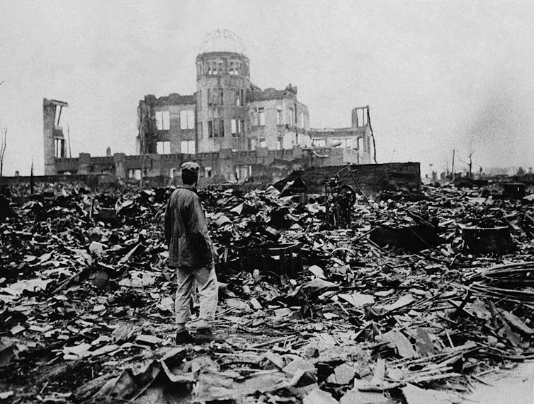 A black and white photo shows a man standing in a sea of rubble, with the ruins of one building in the background.