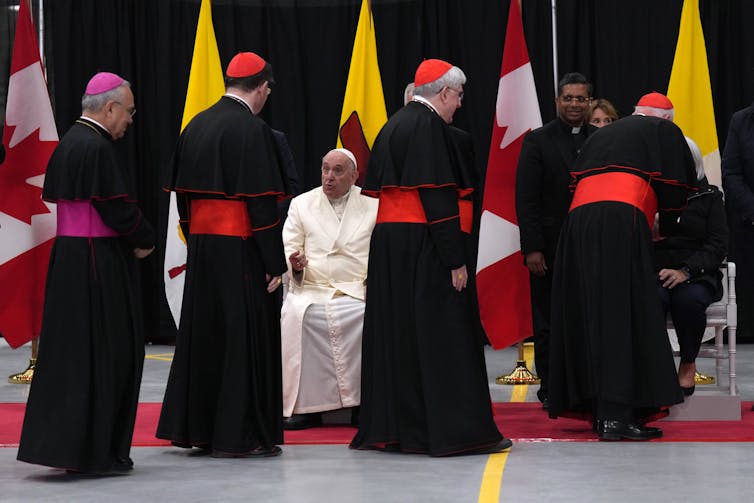 Men in black clerical robes are seen walking past a seated man in a white clerical robe.