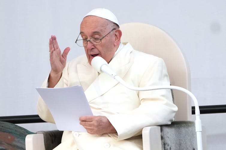 A man in white clerical robes and a skullcap is seen seated and speaking.