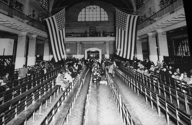 A black and white photo shows a large room with American flags hanging over top and people sitting on wooden benches.