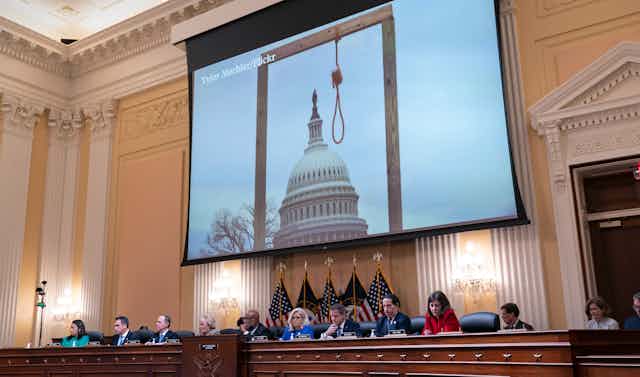 A row of congressional representatives sit in front of a giant screen that shows a gallows and a noose in front of the U.S. Capitol building.