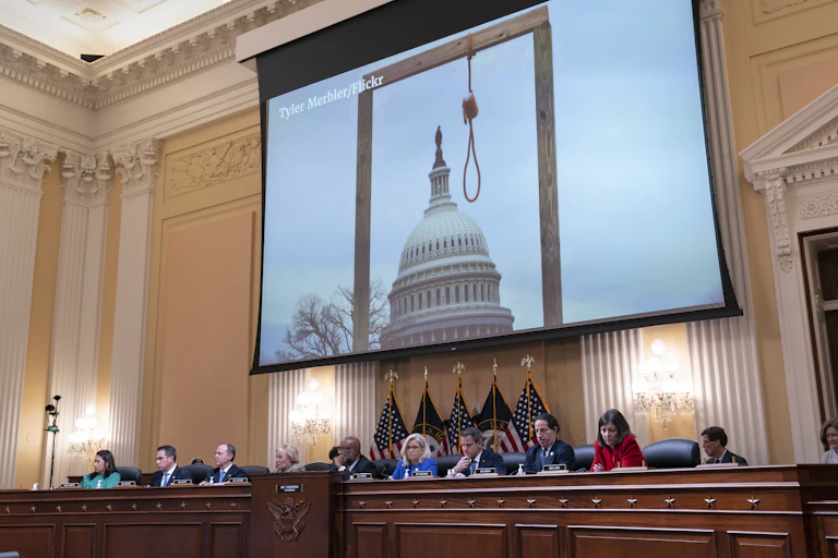 A row of congressional representatives sit in front of a giant screen that shows a gallows and a noose in front of the U.S. Capitol building.