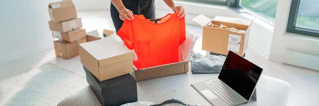 A person opening a package containing an orange t-shirt, unopened parcels are in a pile in the background. 