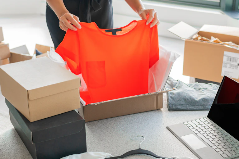 A person opening a package containing an orange t-shirt, unopened parcels are in a pile in the background.