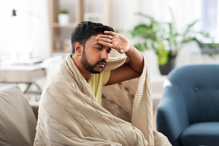 A young man wrapped in a blanket feels his forehead.