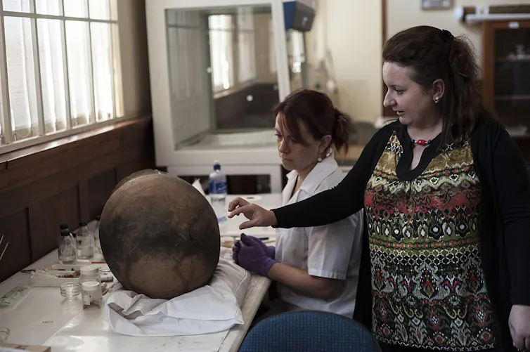 Two women, one seated, examine a clay pot on a table in front of them.