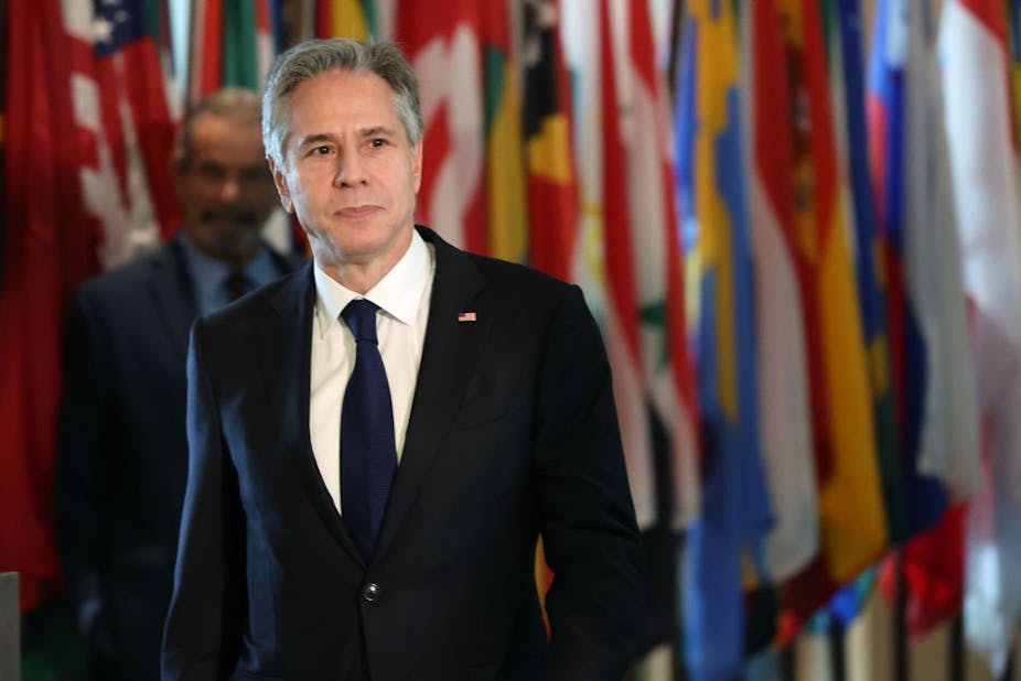 A man in a dark suit and tie, against a background of national flags.