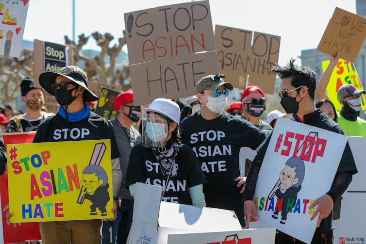 People wear masks and hats and appear to protest in the streets, holding signs that say stop Asian hate.
